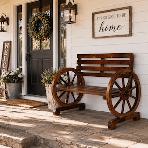 May include: A dark brown wooden bench featuring two large wagon wheels as side supports, a slatted backrest, and a slatted seat. The bench is positioned on a stone porch in front of a white wall with a sign that reads IT'S SO GOOD TO BE home.