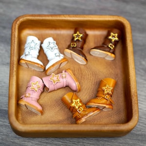 May include: A collection of miniature doll boots in white, dark brown, light pink, and tan colors. Each boot features a gold-toned star emblem on the side, a buckle strap detail, and a light-colored sole. The boots are arranged inside a square wooden bowl.