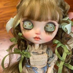 Pu&ograve; includere: Una bambola Blythe personalizzata con capelli castano chiaro acconciati a codini, adornata con nastri verdi. La bambola ha grandi occhi con un motivo floreale e indossa un vestito in stile vintage con pizzo e dettagli blu.
