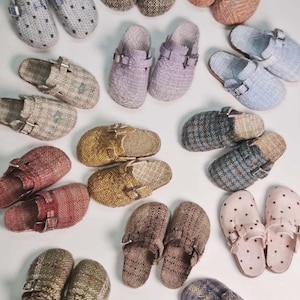 May include: A collection of various slip-on clogs featuring adjustable metal buckles and diverse fabric textures, including woven patterns, solid colors, and polka dots in shades of gray, brown, pink, blue, yellow, and purple arranged on a surface.