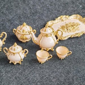May include: A miniature tea set featuring a teapot, two lidded sugar bowls, a creamer, two teacups, and a rectangular serving tray. Each piece is finished in a pale pink color with ornate gold-toned trim and decorative gold accents.