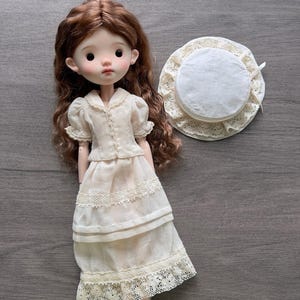 May include: A ball-jointed doll with long wavy brown hair, wearing a cream-coloured short-sleeved dress with lace trim and a tiered skirt. Beside the doll is a matching cream-coloured round hat with a wide lace-trimmed brim and a small ribbon detail.