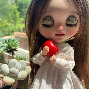 Pu&ograve; includere: Una bambola Blythe con lunghi capelli castani e grandi occhi, indossa un vestito di pizzo bianco, tiene in mano una ciliegia rosso vivo. La bambola &egrave; posizionata davanti a uno sfondo verde sfocato, con una pianta succulenta visibile a sinistra.