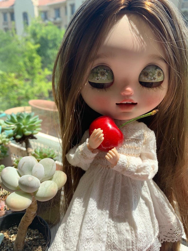 Pu&ograve; includere: Una bambola Blythe con lunghi capelli castani e grandi occhi, indossa un vestito di pizzo bianco, tiene in mano una ciliegia rosso vivo. La bambola &egrave; posizionata davanti a uno sfondo verde sfocato, con una pianta succulenta visibile a sinistra.