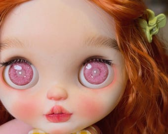 Puede incluir: Una muñeca con cabello naranja vibrante, piel pálida y ojos grandes, redondos y rosados con iris detallados. La muñeca presenta un rubor rosado suave en las mejillas, cejas definidas y una pequeña pinza para el cabello verde claro.