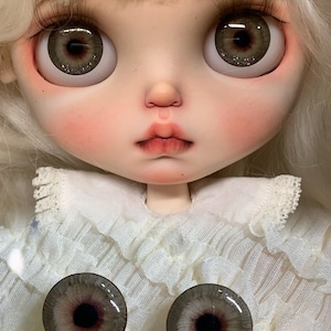 May include: A doll with pale skin, long white hair, and large grey eyes with glitter accents. The doll has a small nose, pink-tinted lips, and wears a white ruffled collar shirt. Two additional circular grey eye chips with glitter are placed below.