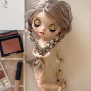 Pu&ograve; includere: Una bambola personalizzata con capelli grigi intrecciati e grandi occhi espressivi. La bambola &egrave; nuda e posa con una gamba piegata. Trucco e accessori sono visibili sullo sfondo, suggerendo un focus sulla bellezza e sull'arte.