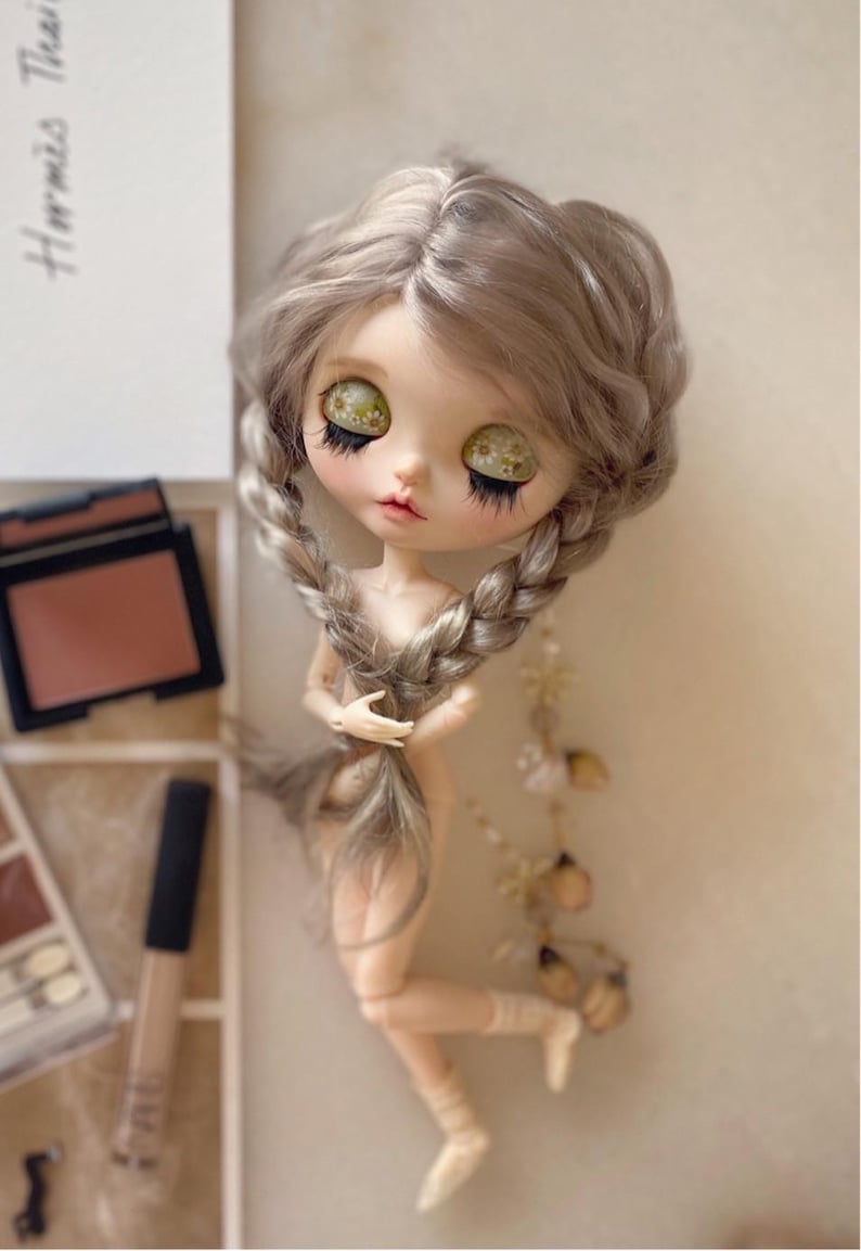 Pu&ograve; includere: Una bambola personalizzata con capelli grigi intrecciati e grandi occhi espressivi. La bambola &egrave; nuda e posa con una gamba piegata. Trucco e accessori sono visibili sullo sfondo, suggerendo un focus sulla bellezza e sull'arte.