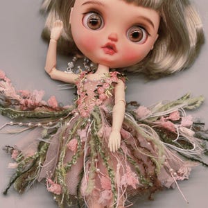 Può includere: Una bambola con capelli corti castano chiaro e grandi occhi marroni indossa un vestito decorato con ricami floreali rosa, accenti di filato verde e perline bianche. Il testo "the TASTE of THINGS" e "(In Winter)" appare in alto.