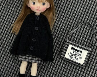 Può includere: Una bambola con lunghi capelli castano chiaro indossa un cardigan nero a trecce, una gonna a quadri bianchi e neri, calzini neri e scarpe nere. Accanto alla bambola c'è una piccola borsa a quadri con una toppa bianca raffigurante un cucciolo e la scritta puppy.
