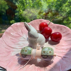 Pu&ograve; includere: Una statuina di coniglio in ceramica bianca e tre ciliegie rosse poggiano su un piatto rosa a forma di foglia. Due piccoli oggetti decorativi a forma di occhio con disegni floreali e ciglia sono anche sul piatto.