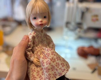 May include: A small doll with blonde hair and bangs, wearing a cream-colored dress featuring a repeating pattern of small red flowers and vines. The doll has dark eyes, rosy cheeks, and is wearing dark brown lace-up boots on its feet.