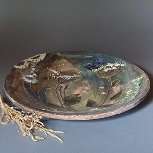Può includere: Un piatto rotondo in ceramica con una superficie ruvida nei toni della terra, marrone e blu. Il piatto presenta un disegno dipinto di piante con fiori bianchi, piccoli fiori rosa e una farfalla blu su un fiore, con un mazzetto di piante essiccate.