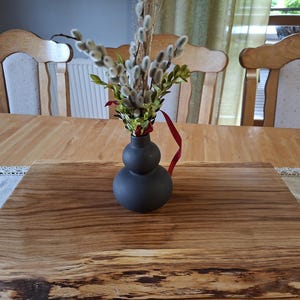 Può includere: Un vaso in ceramica nero opaco a forma di doppio bulbo contenente un bouquet di rami di salice e fogliame verde, legato con un sottile nastro rosso, poggiato su un tagliere rettangolare in legno con bordo naturale grezzo.