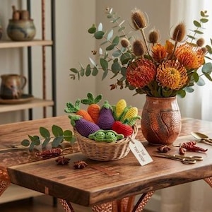 May include: A small woven basket filled with crocheted vegetables including carrots, eggplants, and peas, accompanied by a vase of orange pincushion flowers and eucalyptus, all resting on a wooden table with copper-toned metal legs.
