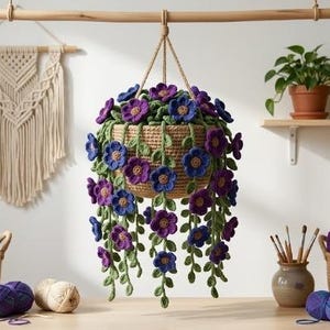 May include: A crocheted hanging plant in a tan basket featuring purple and blue flowers with green stems and leaves. The plant is suspended from a wooden rod. The scene includes various balls of yarn in baskets and a macrame wall hanging.