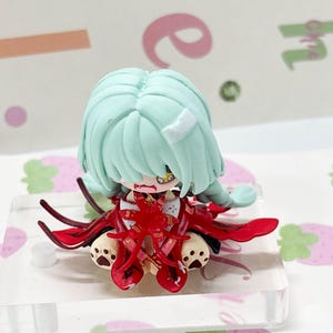 May include: A small chibi-style figurine with mint green hair, a white bandage on the head, and a red outfit with black accents. The character has a yellow eye, visible fangs, and sits on a clear acrylic base with paw-print details on the feet.