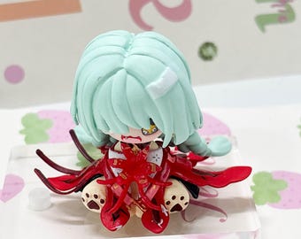 May include: A small chibi-style figurine with mint green hair, a white bandage on the head, and a red outfit with black accents. The character has a yellow eye, visible fangs, and sits on a clear acrylic base with paw-print details on the feet.