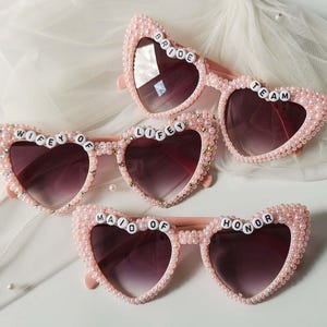 Op de afbeelding: Vier paar roze hartvormige zonnebrillen met parel- en strassversieringen. De zonnebrillen hebben donkere glazen en de tekst "BRIDE", "TEAM", "WIFEY", "LIFEY" en "MAID OF HONOR".