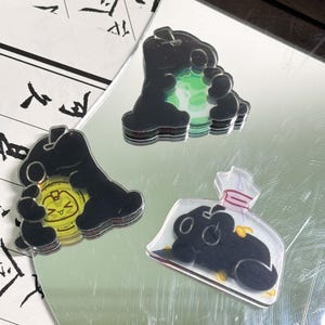 以下が含まれることがあります： 黒い丸みを帯びた生き物の形をしたアクリルチャーム3点。1つは光る緑色の球体を持ち、もう1つは顔が描かれた黄色いコインを抱きしめ、3つ目はピンク色の栓が付いた透明なボトル型の容器の中に入っています。
