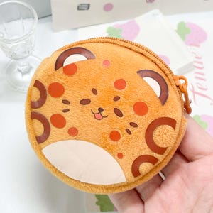 May include: A round, plush, orange coin purse featuring a printed leopard-like face with dark brown spots, a small smiling mouth, and a cream-colored chin area. The pouch has a matching orange zipper closure along the top edge.