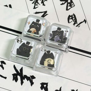 May include: Four square, clear plastic mechanical keyboard keycaps featuring illustrations of black cats wearing top hats. Each keycap displays a unique cat graphic, including one with a gold coin, one with a dollar sign, and Chinese characters.
