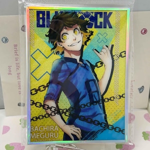 Könnte beinhalten: Eine rechteckige holografische Sammelkarte mit einer Anime-Figur mit dunklem Haar und gelben Augen in einem blauen Trikot. Der Hintergrund ist gelb mit blauem Text BLUE LOCK und BACHIRA MEGURU, überlagert mit schwarzen Kettengrafiken.
