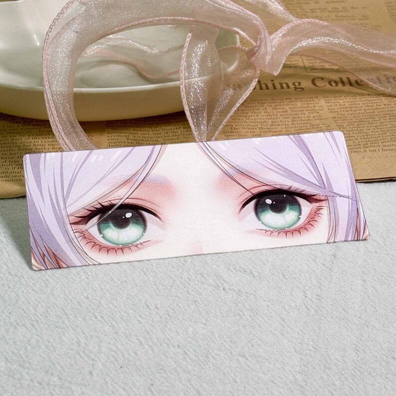 K&ouml;nnte beinhalten: Ein rechteckiges Lesezeichen mit einer detaillierten Anime-Illustration eines Paares gro&szlig;er, ausdrucksstarker Augen mit hellgr&uuml;nen Iris und langen Wimpern, umrahmt von blasslavendelfarbenem Haar, befestigt an einem hellrosa Organzaband.