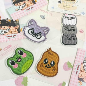 May include: A collection of four embroidered patches featuring a purple wolf head with glasses, a stack of three cats, a green frog with pink cheeks, and a brown textured character, all displayed on individual backing cards with illustrated designs.
