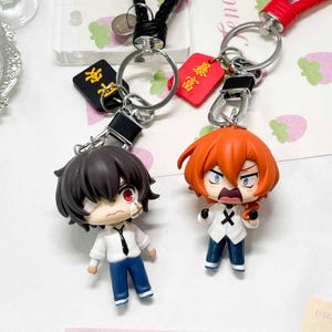 Könnte beinhalten: Zwei Schlüsselanhänger im Anime-Stil mit Metallringen und geflochtenen Bändern. Eine Figur hat dunkles Haar, ein weißes Hemd und eine blaue Hose. Die andere hat orangefarbenes Haar, ein weißes Hemd mit einem schwarzen X und eine blaue Hose. Beide haben kleine rechteckige Anhänger mit chinesischen Schriftzeichen.