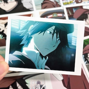 Könnte beinhalten: Ein rechteckiger Fotoabzug mit einer Anime-Illustration einer Figur mit dunklem Haar und einer Mütze. Die Figur ist im Profil mit ernstem Gesichtsausdruck dargestellt und in kühlen Blau- und Türkistönen mit hellen Lichtakzenten gehalten.