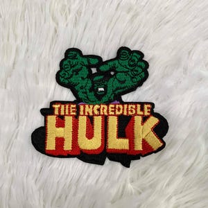 Może przedstawiać: Haftowana naszywka z Incredible Hulk. Projekt zawiera zielone dłonie i twarz Hulka nad napisem "THE INCREDIBLE HULK" w czerwonych i złotych literach, obrysowanych na czarno. Naszywka jest prawdopodobnie przeznaczona na odzież lub akcesoria.