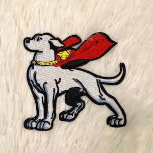 May include: Embroidered patch featuring a white dog wearing a red cape with a yellow Superman logo. The dog has a yellow collar and black accents. The patch is outlined in black and blue.