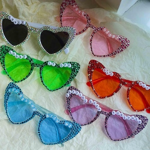 May include: Several pairs of heart-shaped sunglasses in shades of pink, green, blue, and red. Each pair is decorated with rhinestones and has text like "STYLE", "LOVE", "SHAKE IT OFF", "BAD BLOOD", and "GETTING BACK TOGETHER".