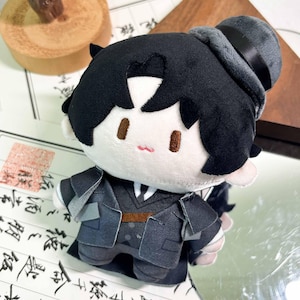 May include: A plush doll featuring a character with black hair, wearing a dark gray suit, a white shirt with a black tie, and a dark gray top hat. The doll has embroidered brown eyes and a small pink mouth, with a brown belt detail on the suit.