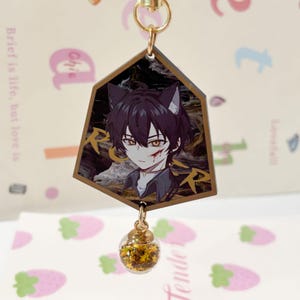 Könnte beinhalten: Ein fünfeckiger Acryl-Schlüsselanhänger mit einer Anime-Figur mit dunklem Haar, Katzenohren und einem roten Fleck auf der Wange. Der Hintergrund ist ein dunkles, wirbelndes Muster mit goldener Schrift. Ein kleiner, durchsichtiger, kugelförmiger Anhänger, gefüllt mit goldenem Sternenglitzer, hängt an der Unterseite.