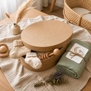 May include: A baby gift set in an oval cardboard box, including a green blanket, wooden rattle, and knitted socks. A small vase with dried flowers and a woven bassinet are in the background. The blanket has a card tied with twine.