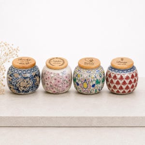 May include: Four round ceramic jars with wooden lids, each featuring unique patterns: blue and white floral, pink cherry blossoms, colorful botanical motifs, and red geometric designs. Each lid is engraved with a name and date.