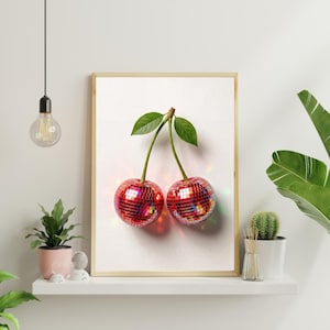 May include: A framed print featuring two cherries with red, mirrored disco ball surfaces, green stems, and leaves. The artwork is set against a white background, with a light wooden frame. The image is displayed on a white shelf with plants.