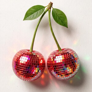 May include: Two red disco ball cherries with green stems and leaves against a white background. The cherries are made of small, reflective squares, creating a sparkling effect. The image has a playful and festive vibe.