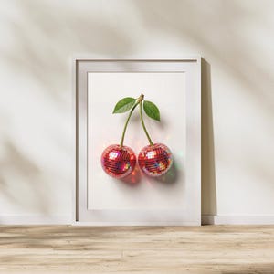 May include: A framed print featuring two cherries with disco ball-like surfaces. The cherries are red with reflective facets, connected by a brown stem and green leaves. The artwork is set against a white background, framed in a white border.