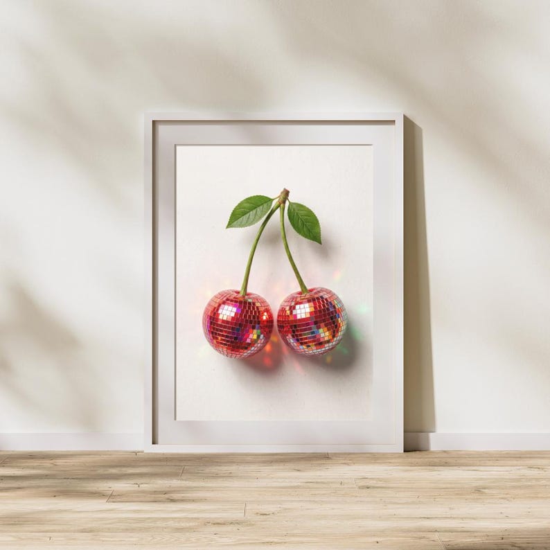 May include: A framed print featuring two cherries with disco ball-like surfaces. The cherries are red with reflective facets, connected by a brown stem and green leaves. The artwork is set against a white background, framed in a white border.