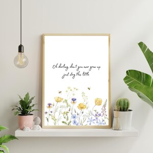 May include: A rectangular wall art print in a light wood frame featuring the black cursive text Oh darling, don't you ever grow up just stay this little above a watercolor illustration of yellow and purple wildflowers with two small bees.