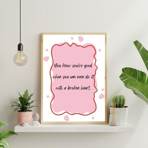 Op de afbeelding: Een rechthoekige wandkunstprint in een lichthouten lijst met een roze achtergrond, een golvende donkerrode rand en de zwarte tekst: You know you're good when you can even do it with a broken heart. Het ontwerp bevat verspreide roze stippen.