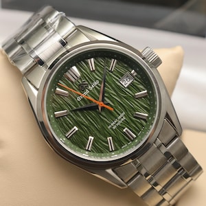 May include: A Grand Seiko wristwatch with a textured green dial, silver-tone hour markers, and a date window at the three o'clock position. The watch features a stainless steel link bracelet, a bright orange second hand, and black hour and minute hands.