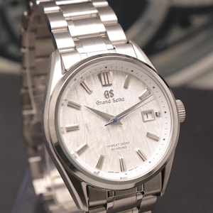 May include: A silver-toned stainless steel wristwatch featuring a textured white dial with silver indices, a date window at the three o'clock position, and the text Grand Seiko, GS, Hi-Beat 36000, and 80 Hours printed on the watch face.