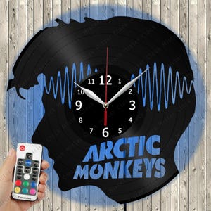 Könnte beinhalten: Eine Wanduhr aus einer schwarzen Schallplatte mit einer blauen Schallwellengrafik und dem Schriftzug Arctic Monkeys. Die Uhr hat weiße Stunden- und Minutenzeiger, einen roten Sekundenzeiger und eine Fernbedienung für LED-Licht.