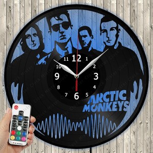 Könnte beinhalten: Eine runde Wanduhr aus einer schwarzen Schallplatte mit einer blauen Grafik der Band Arctic Monkeys, inklusive Bandname und Schallwellenmuster. Die Uhr verfügt über weiße Stunden- und Minutenzeiger sowie einen roten Sekundenzeiger.