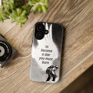 Può includere: Custodia per telefono in bianco e nero con la citazione "to become a star you must burn". La custodia presenta una grafica di una persona con uno zaino. La custodia è su una superficie di legno.