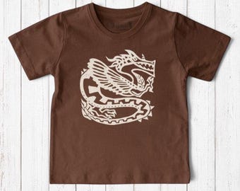 May include: A short-sleeved brown t-shirt featuring a large, cream-colored graphic print of a stylized dragon with wings and a long, coiled tail. The design is centered on the chest, showcasing intricate line work and geometric patterns.